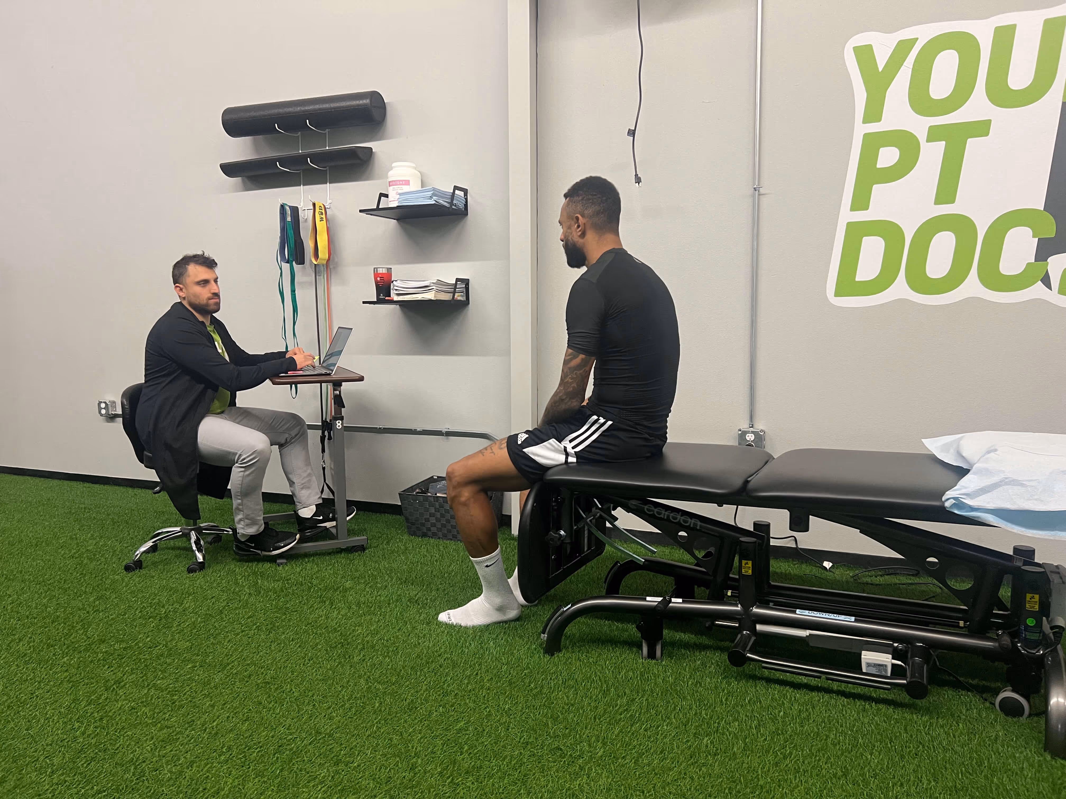 A physical therapist consulting a male athlete sitting on an examination table in a clinic with green artificial turf flooring.
