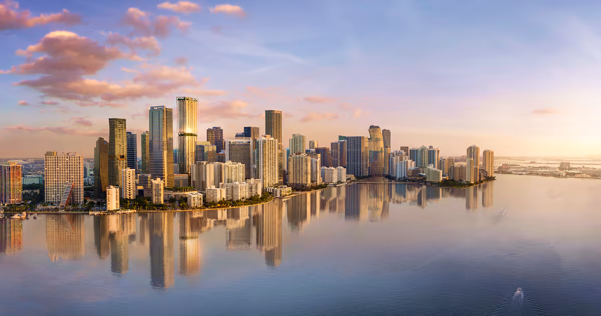 Panoramic view of a city skyline with tall buildings reflected in calm water during a sunset with pink clouds.