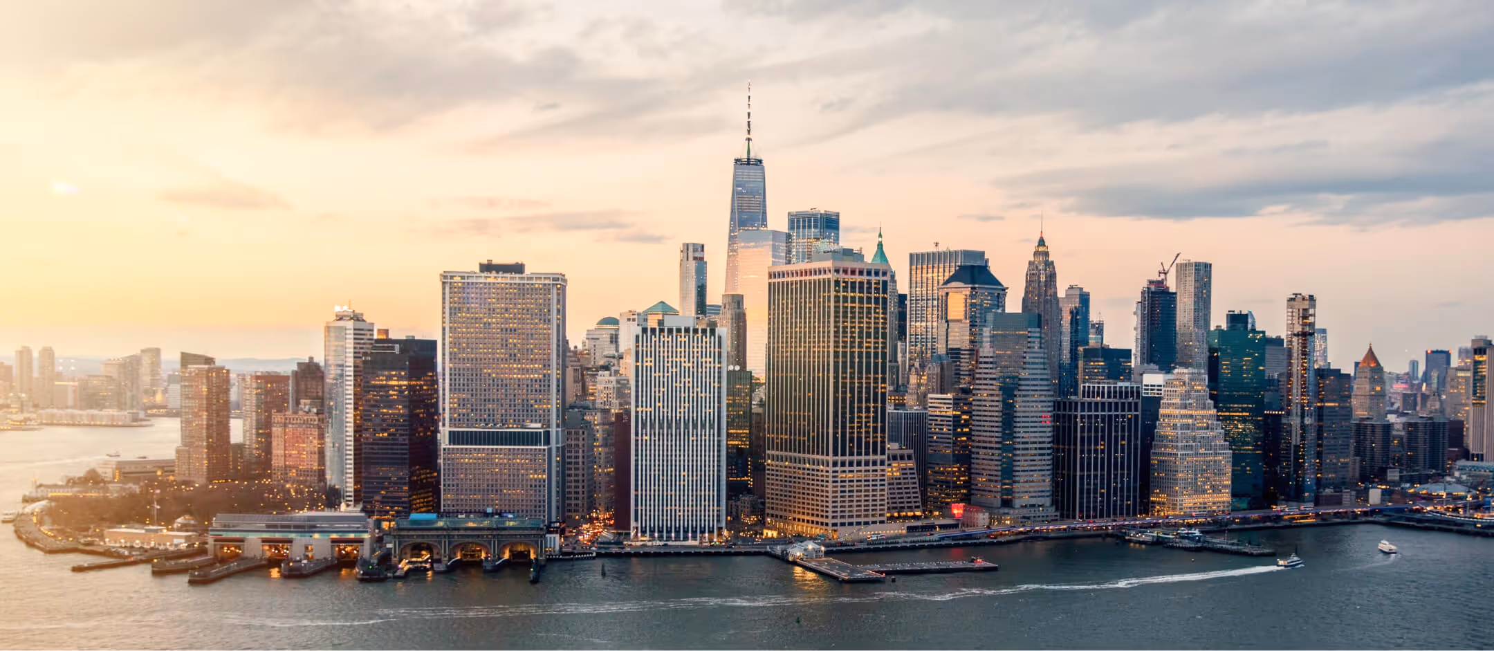 Panoramic view of Lower Manhattan skyline at sunset with One World Trade Center and waterfront piers.