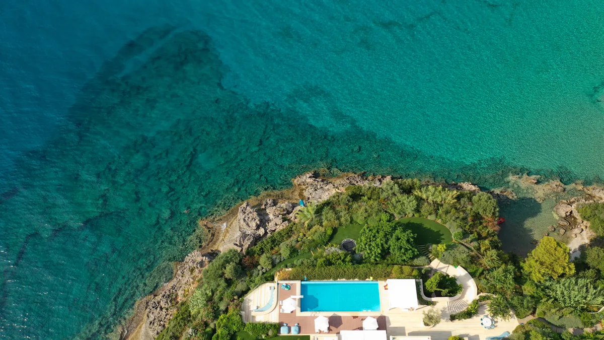 Aerial view of a luxury villa and pool on a green headland above clear turquoise sea