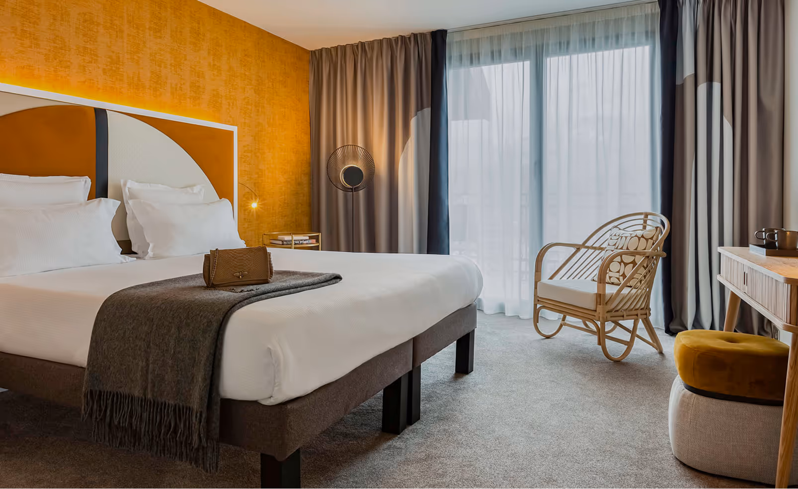 Modern hotel room with a large bed featuring white linens and a gray throw, a brown purse on the bed, a wicker chair with cushion, and floor-to-ceiling windows with sheer curtains.