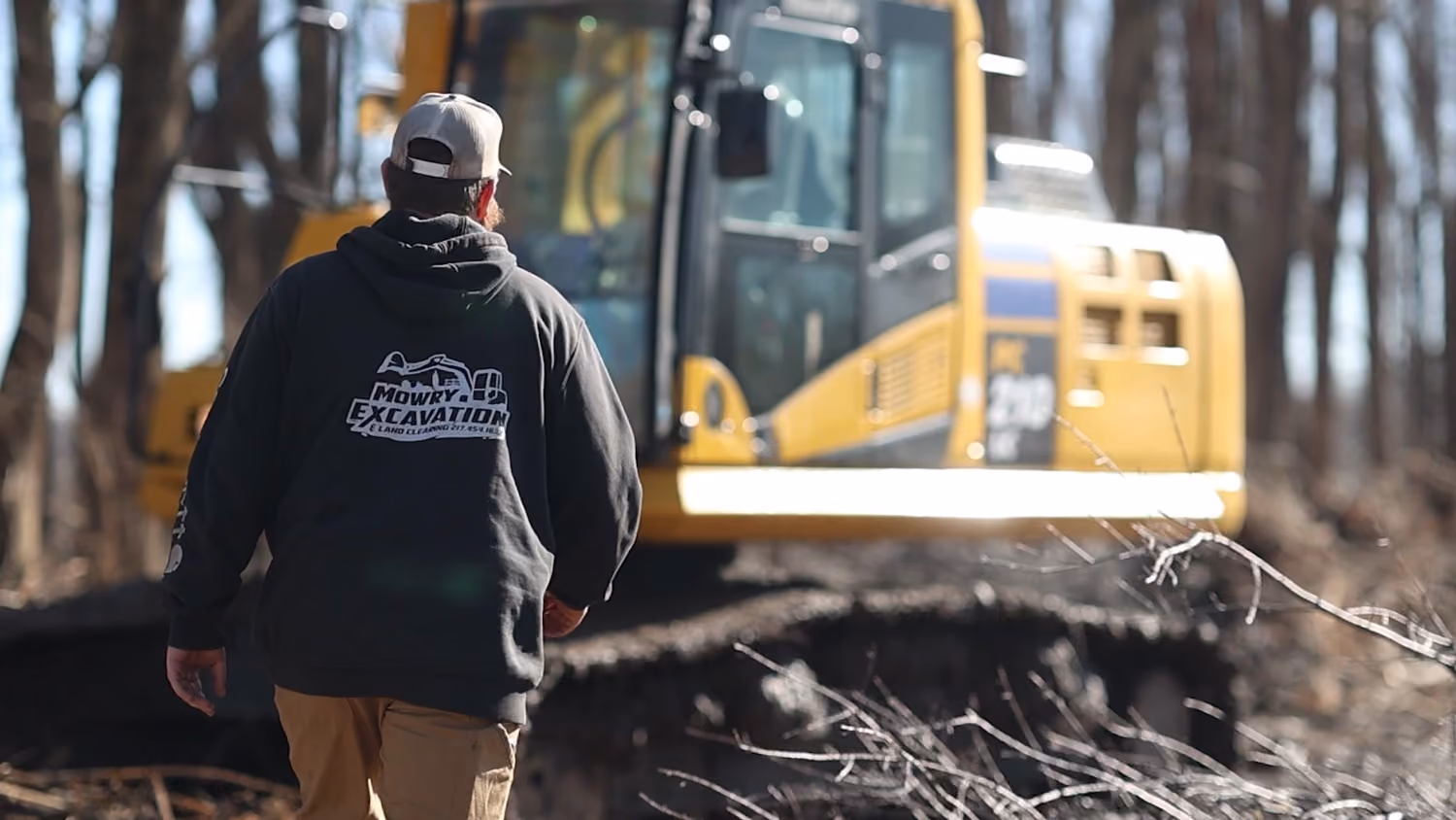 Excavator operator walking toward a yellow excavator on a wooded job site