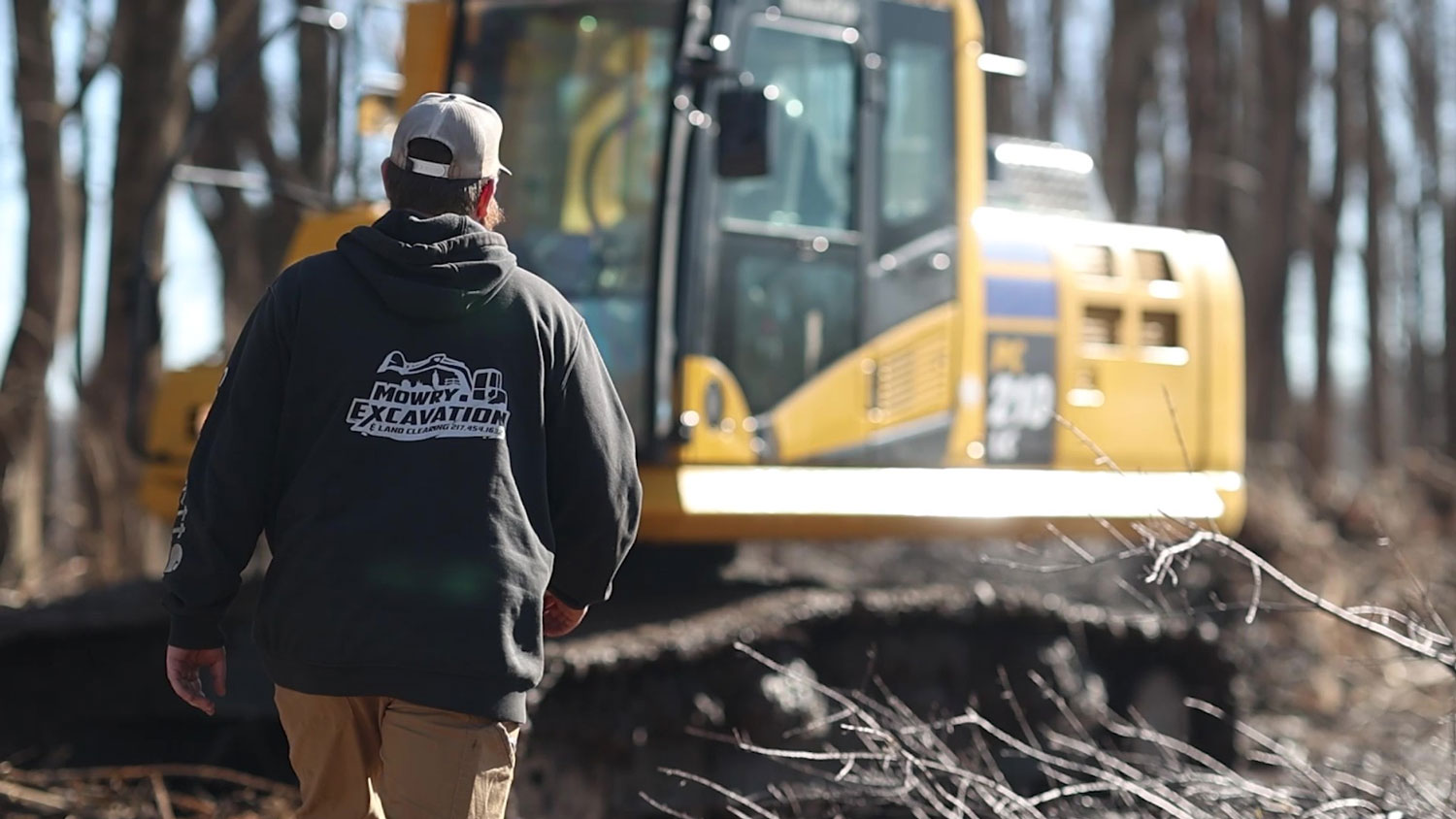 Excavator operator walking toward a yellow excavator on a wooded job site