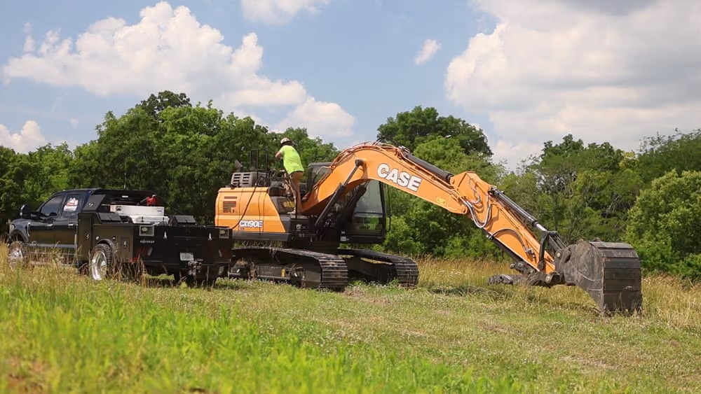 Excavator beside a black pickup truck along a tree line