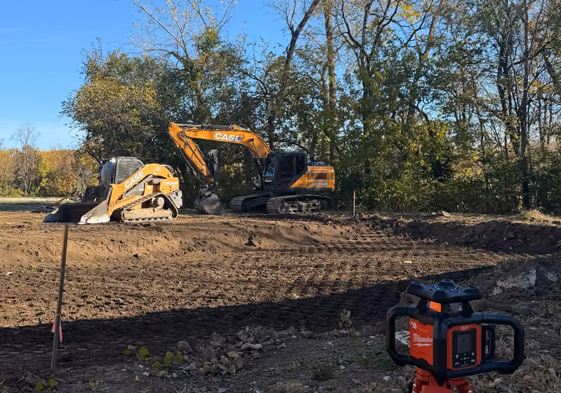 Two excavators on a graded dirt lot with a red rotary laser level in the foreground