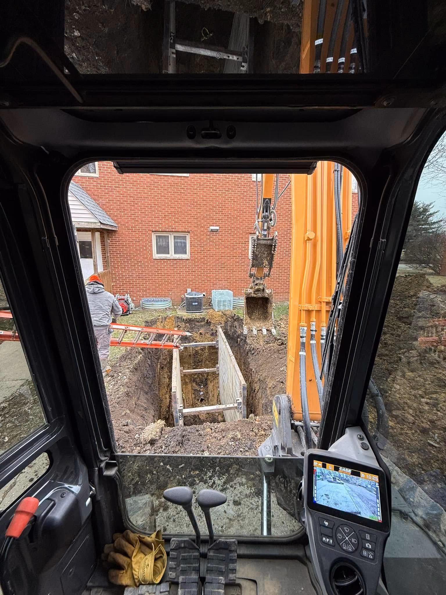 View from an excavator cab of an open utility trench beside a brick building