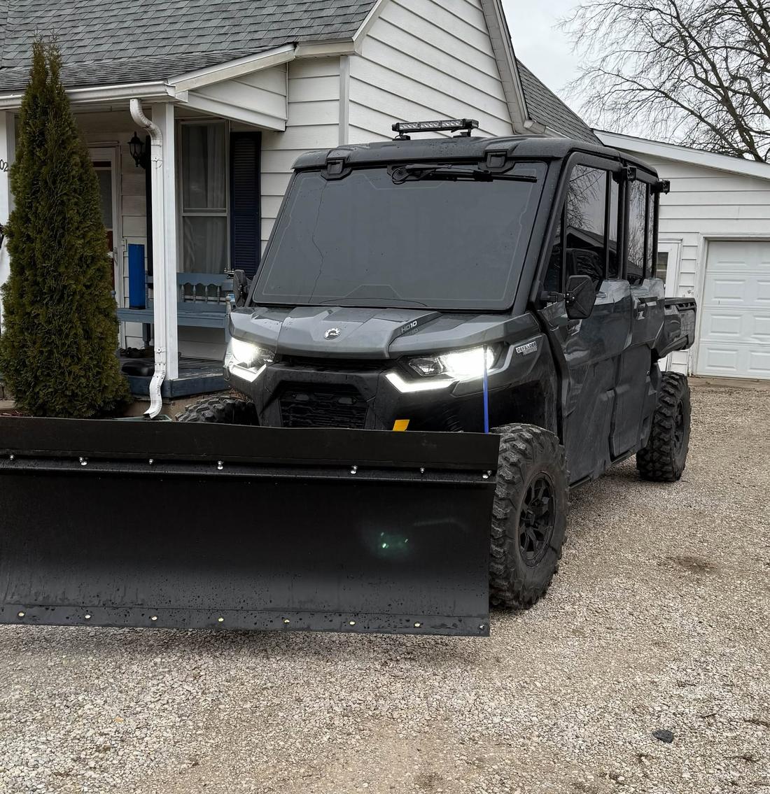 Black side-by-side UTV with a front blade plow on a gravel driveway in front of a white house, Mowry Excavation snow removal equipment