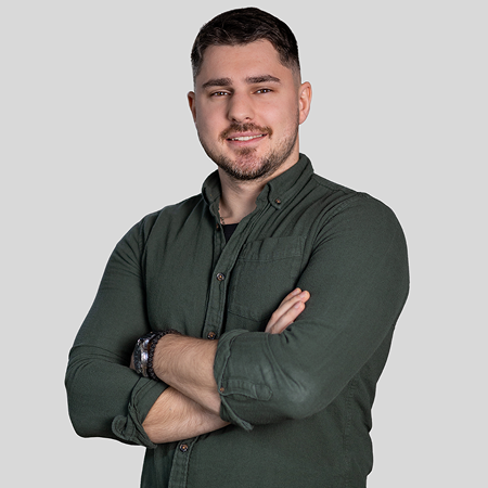 Smiling man with dark hair and beard, wearing a dark green shirt with sleeves rolled up, arms crossed, standing against a gray background.