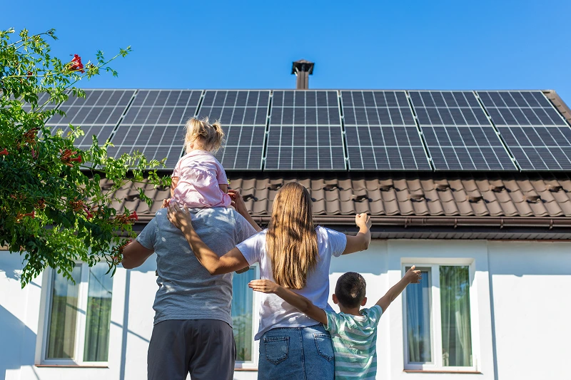 Family of four from behind looking at solar panels installed on the roof of their house under a clear blue sky.