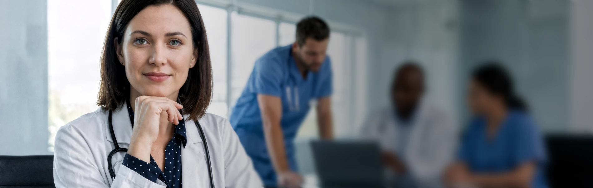 Confident female doctor in white coat with stethoscope looking at camera with medical team working in background.
