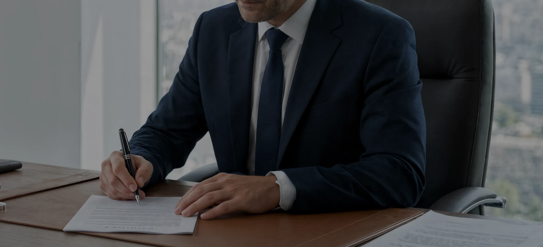 Businessman in a navy suit signing a document at a desk with city view in the background.