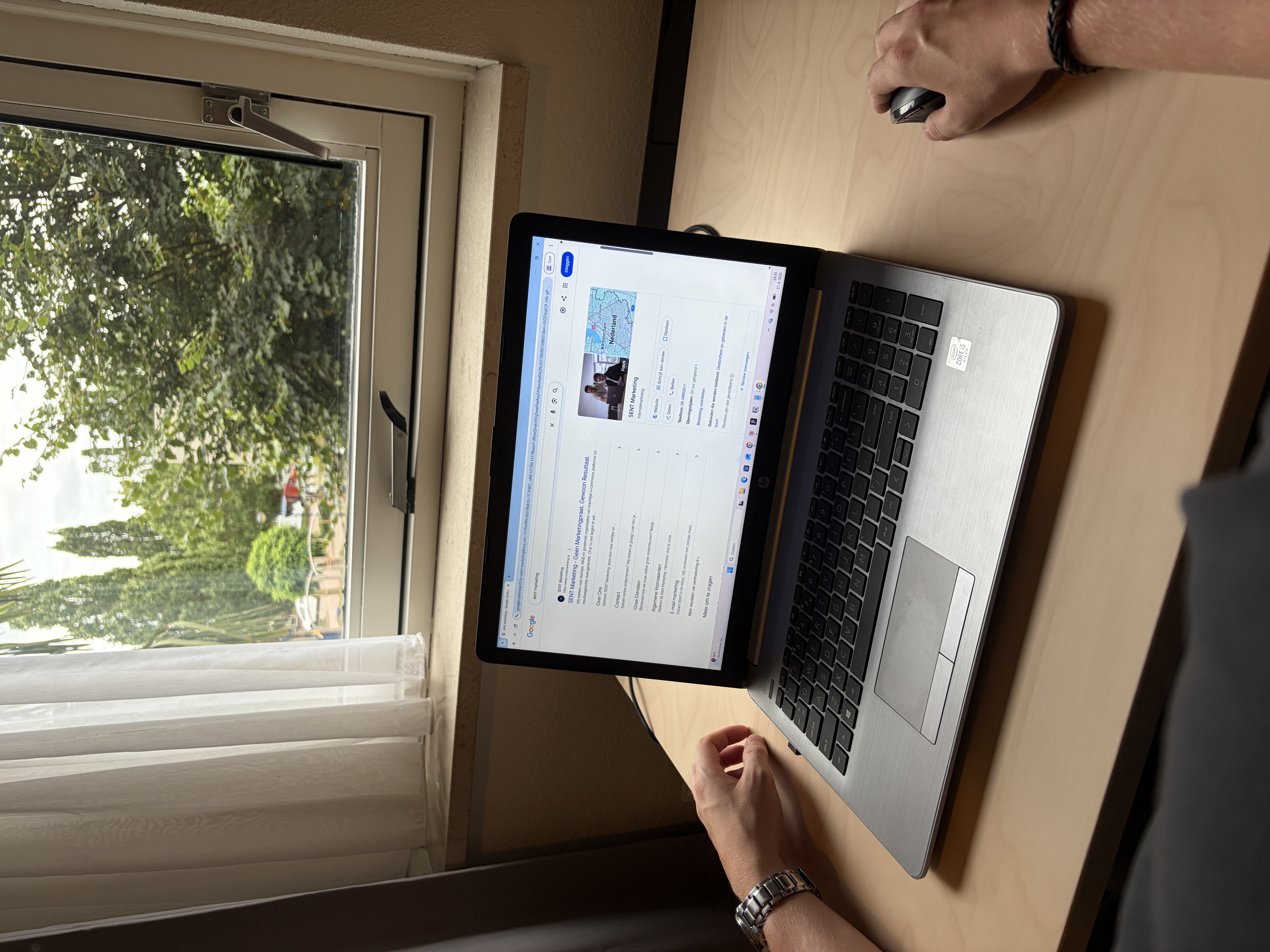 Laptop op een houten bureau bij een raam toont een Google-zoekresultaat voor SENT Marketing, met twee handen ernaast