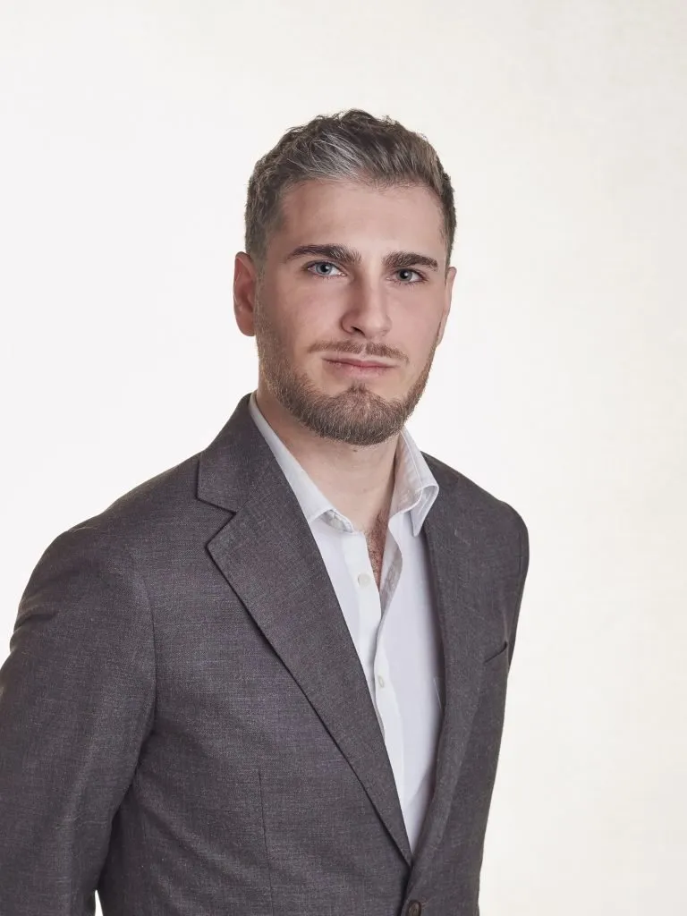 Portrait of a man with short brown hair and beard wearing a gray suit jacket and white shirt, against a light background.