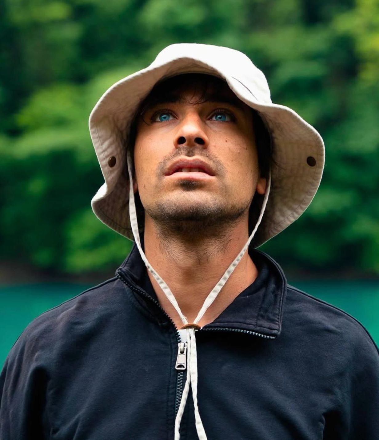Man with blue eyes wearing a white bucket hat and black zip-up jacket looking upward with green foliage background.