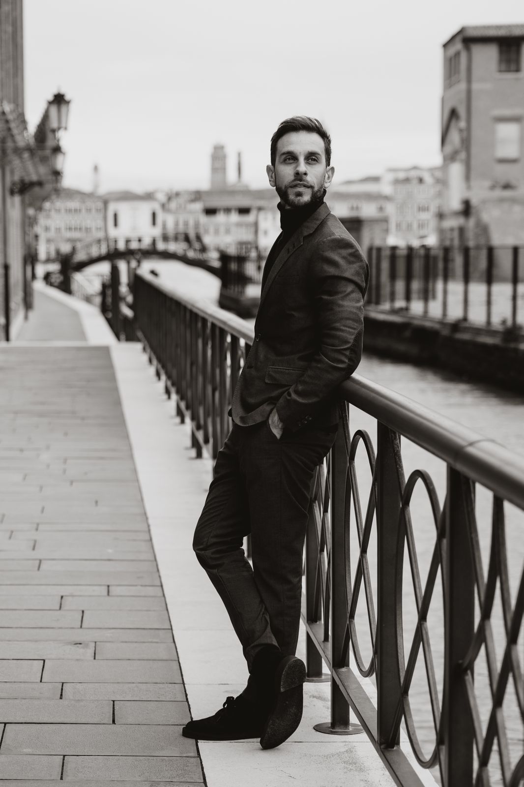 Man in a dark suit leaning against a railing by a canal with a bridge and buildings in the background.