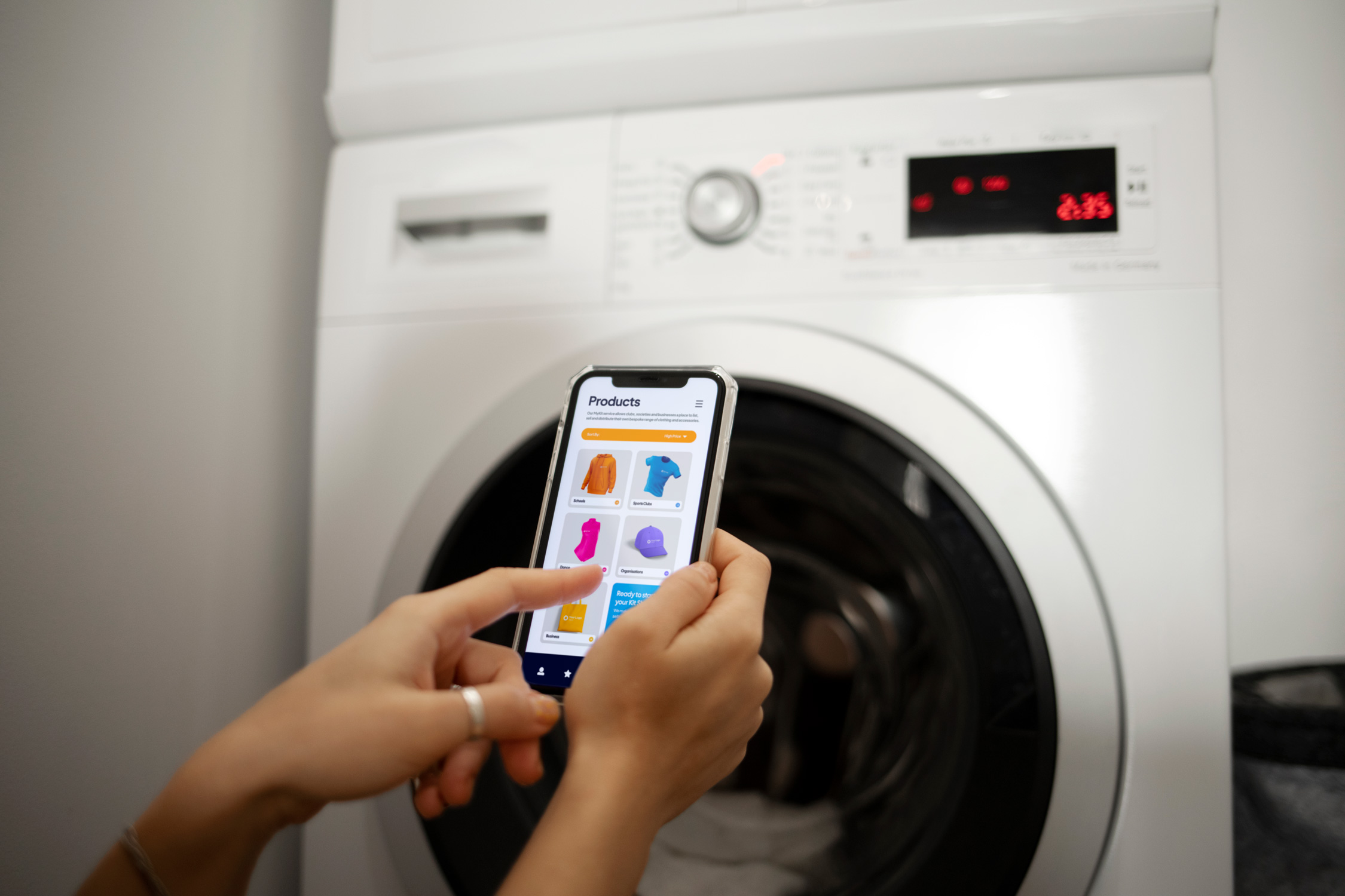 A person uses a smartphone to shop online while standing in front of a washing machine.