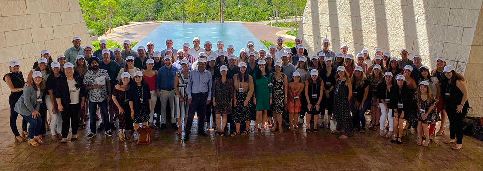 Large diverse group of people wearing white caps standing together under a stone archway with a pool and greenery in the background.