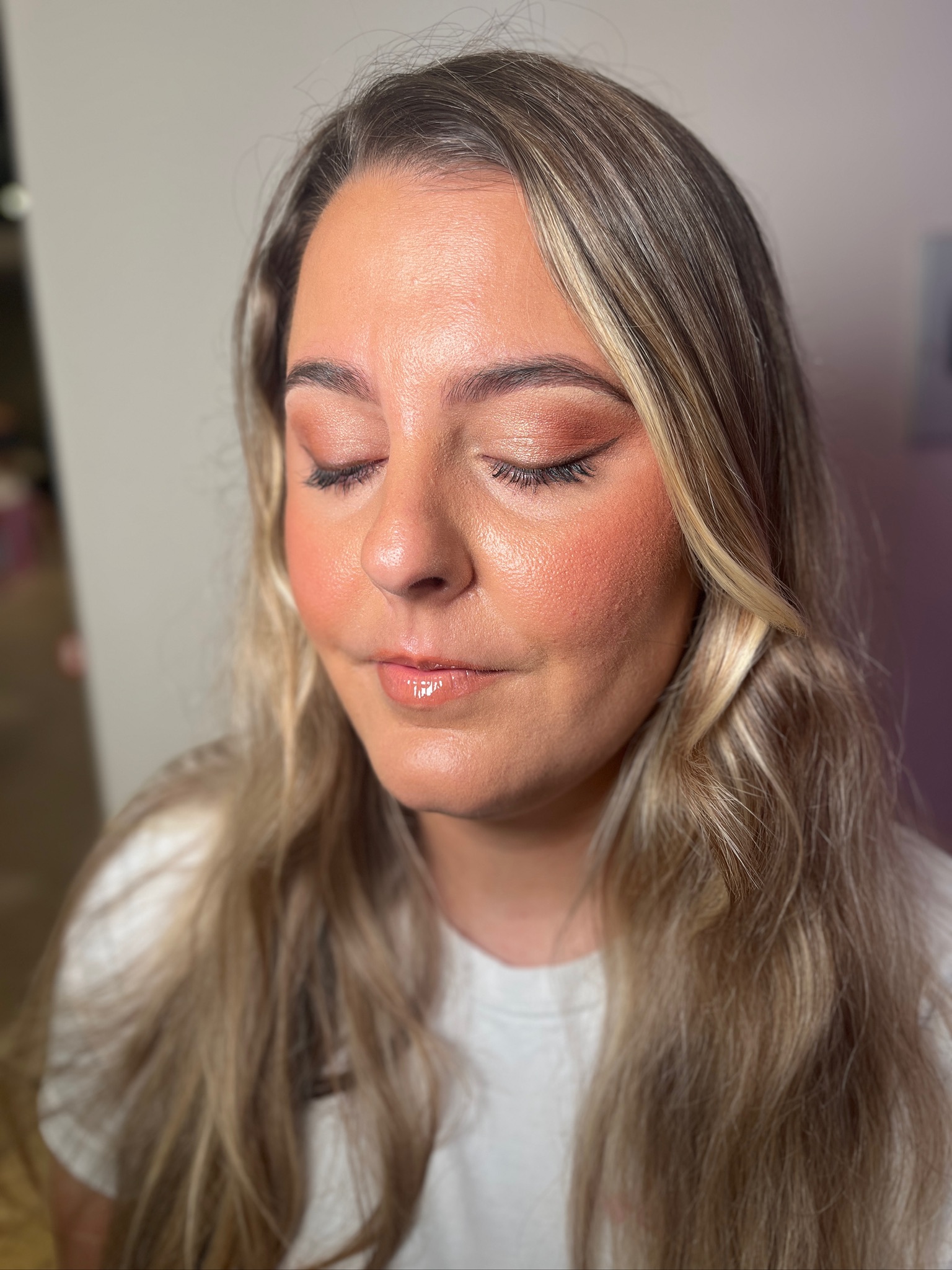 Close-up of a woman with long blonde hair and eyes closed, showing natural makeup with shimmery eyeshadow, mascara, blush, and glossy lips.