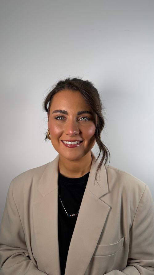 Smiling woman with green eyes and dark hair styled with a loose strand, wearing gold hoop earrings, a beige blazer over a black top, and a necklace with multicolored beads, standing against a plain light gray background.