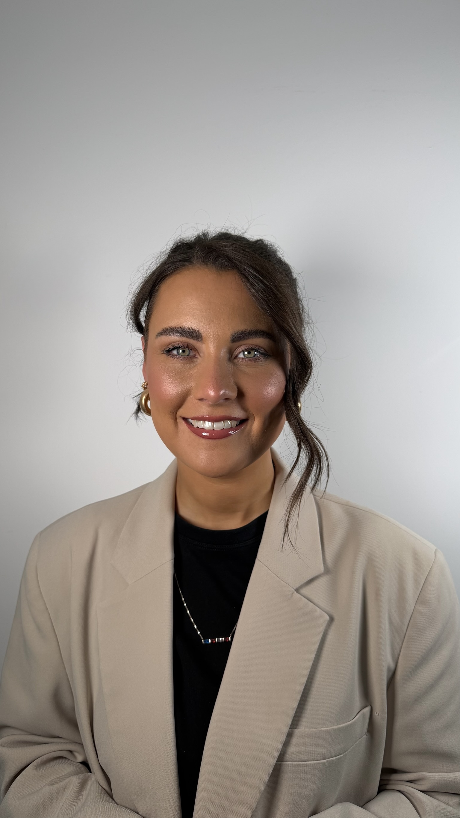 Smiling woman with green eyes and dark hair styled with a loose strand, wearing gold hoop earrings, a beige blazer over a black top, and a necklace with multicolored beads, standing against a plain light gray background.