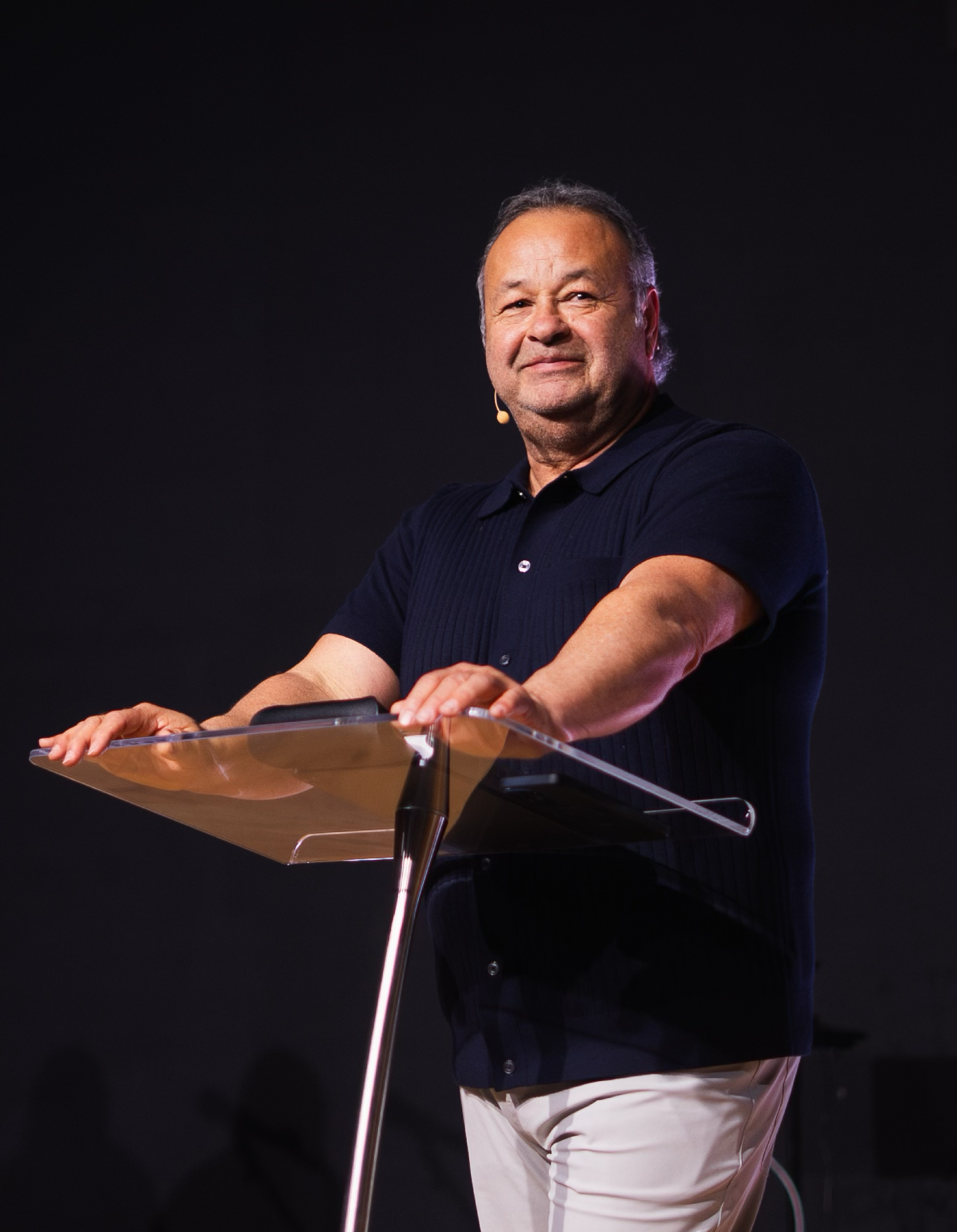 Man wearing a black shirt and beige pants speaking at a clear podium with a microphone headset against a dark background.