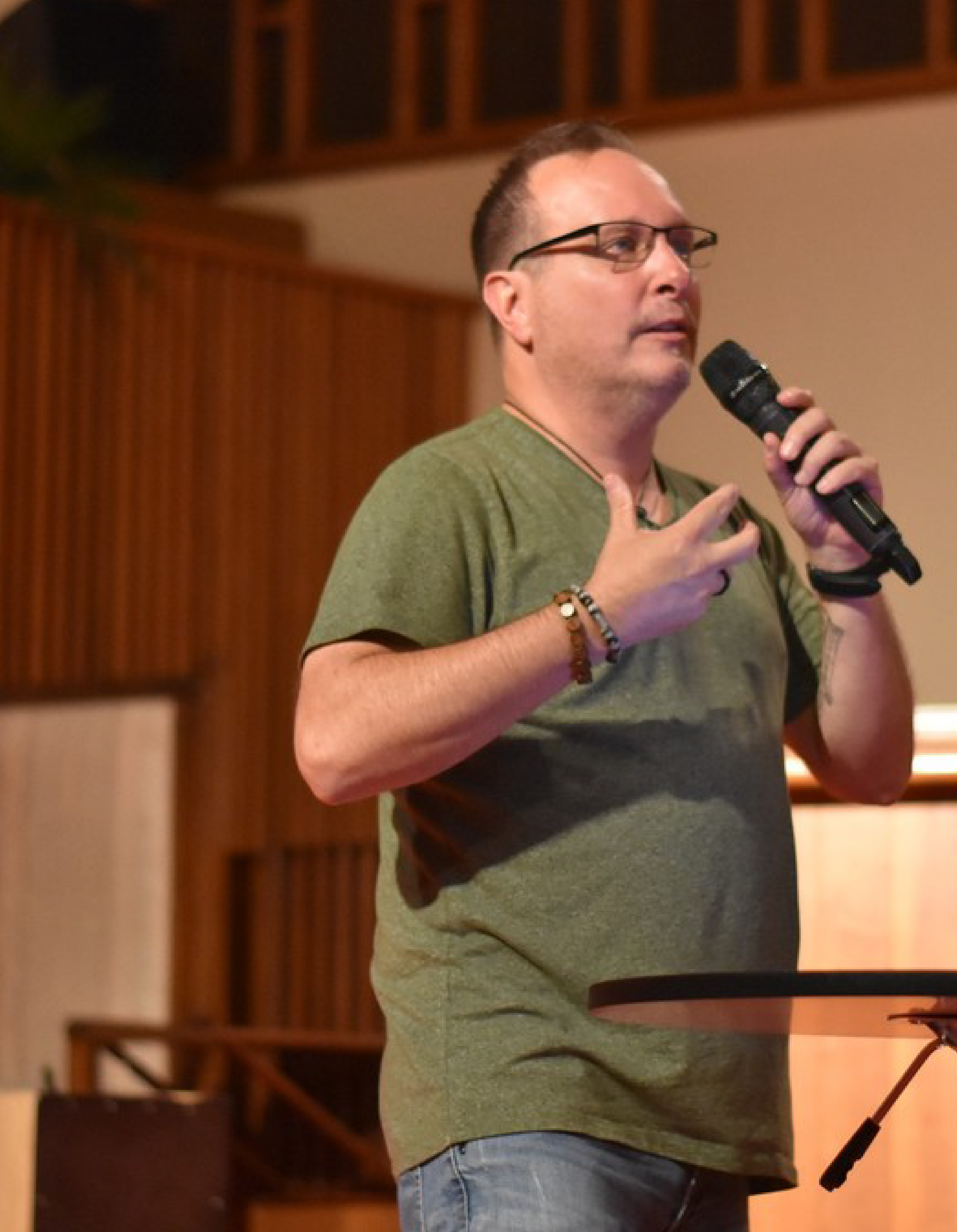 Man in green t-shirt and glasses speaking into a microphone on stage.
