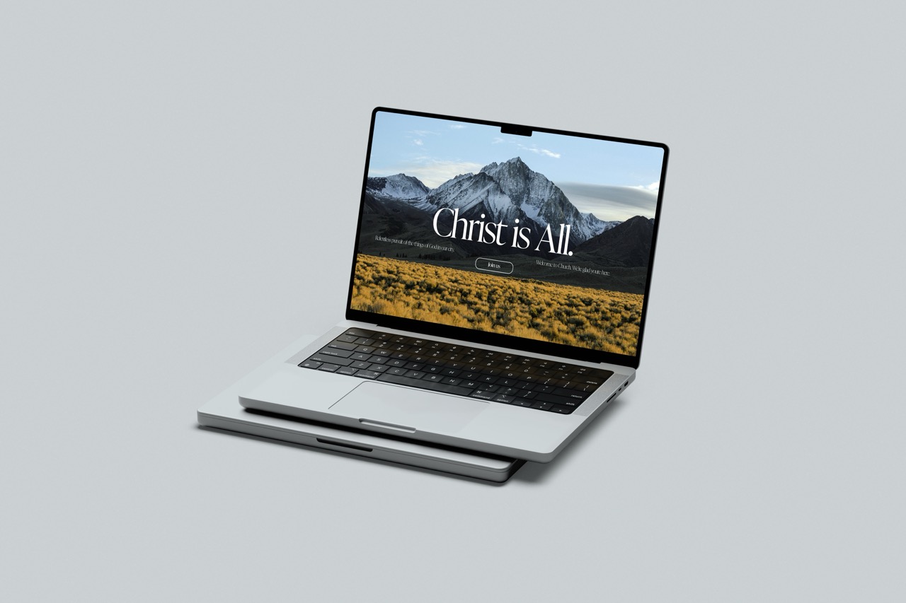 Open laptop displaying a website with a mountain landscape background and the text 'Christ is All.'