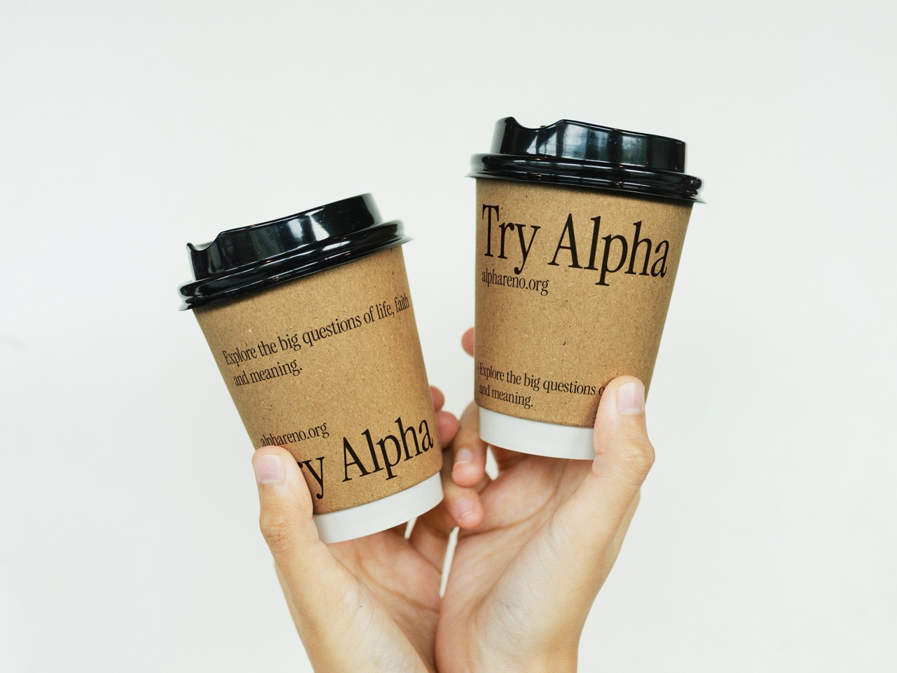 Two hands holding brown disposable coffee cups with black lids, each cup displaying the text 'Try Alpha' and 'Explore the big questions of life, faith and meaning.'