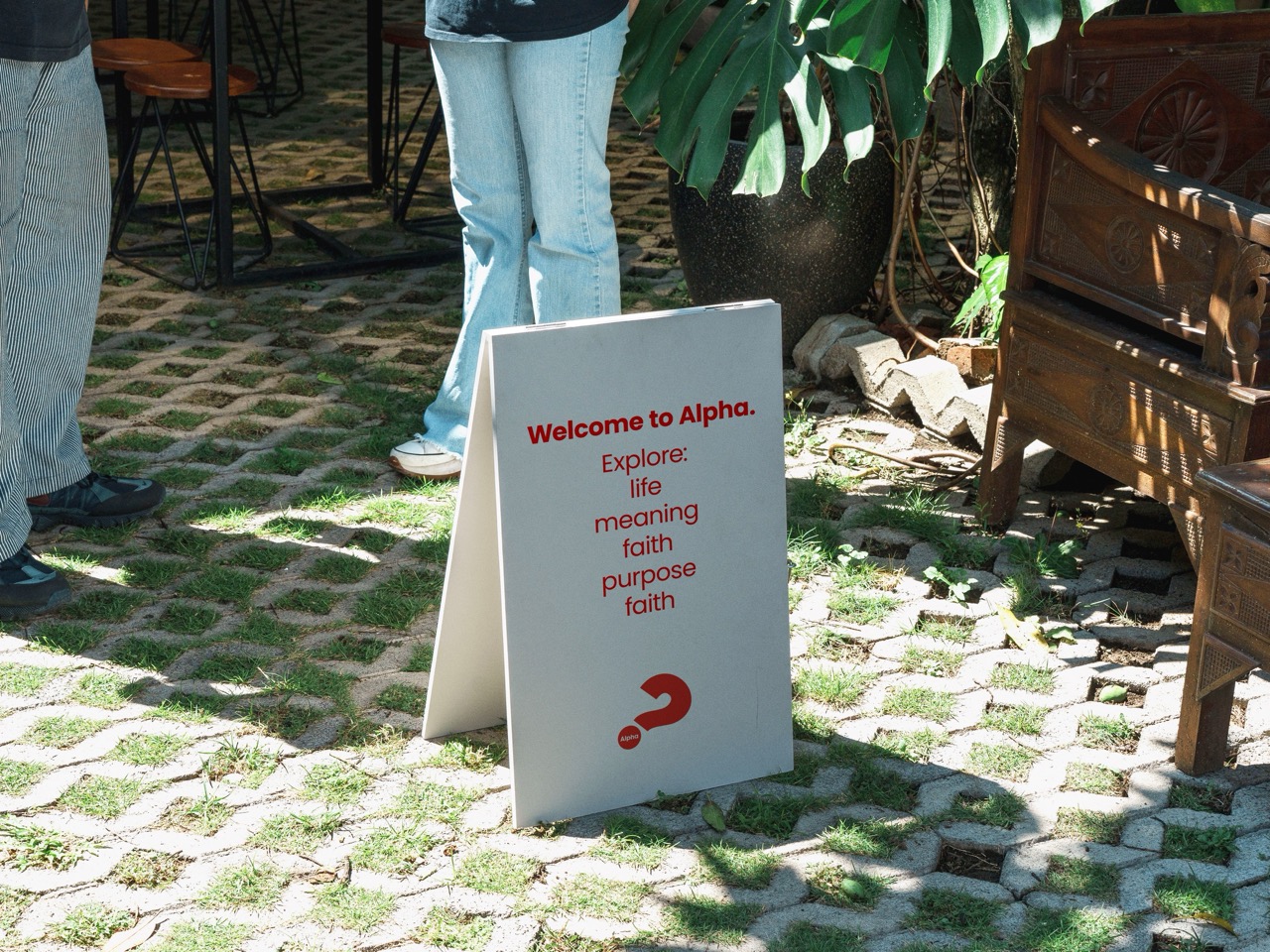 White A-frame sign outdoors that reads 'Welcome to Alpha. Explore: life meaning faith purpose faith' in red text.