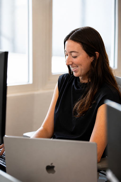 Smiling woman with long brown hair working on a laptop in Nave Bank.
