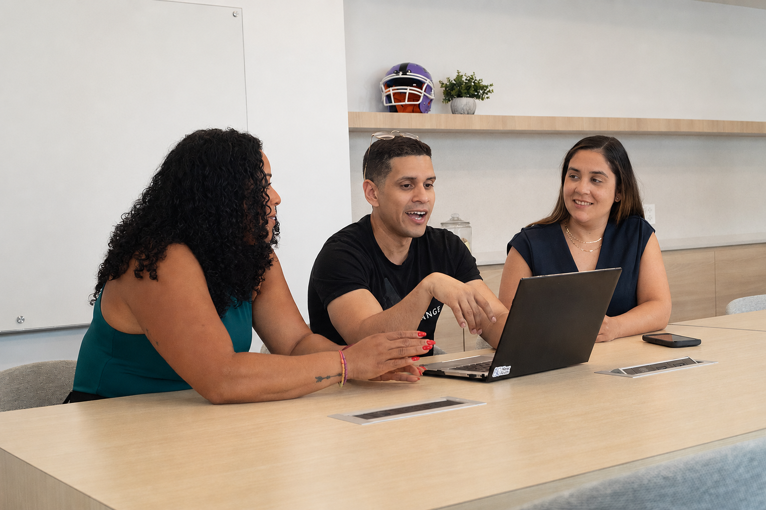 Three people sitting at a table with a laptop, engaged in discussion in a modern office.