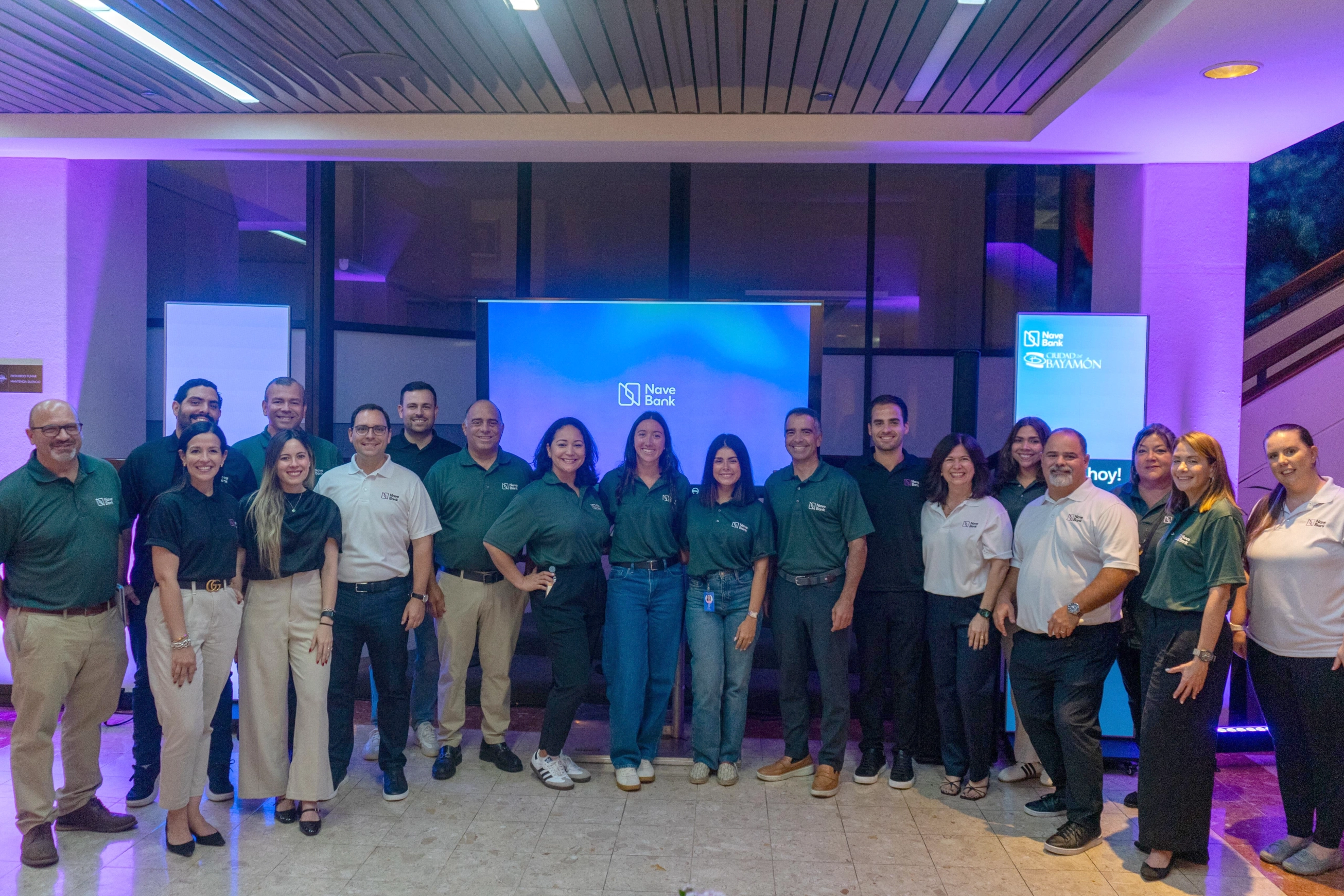 Group of 18 people posing indoors with two blue Nave Bank screens behind them at event in Bayamon.