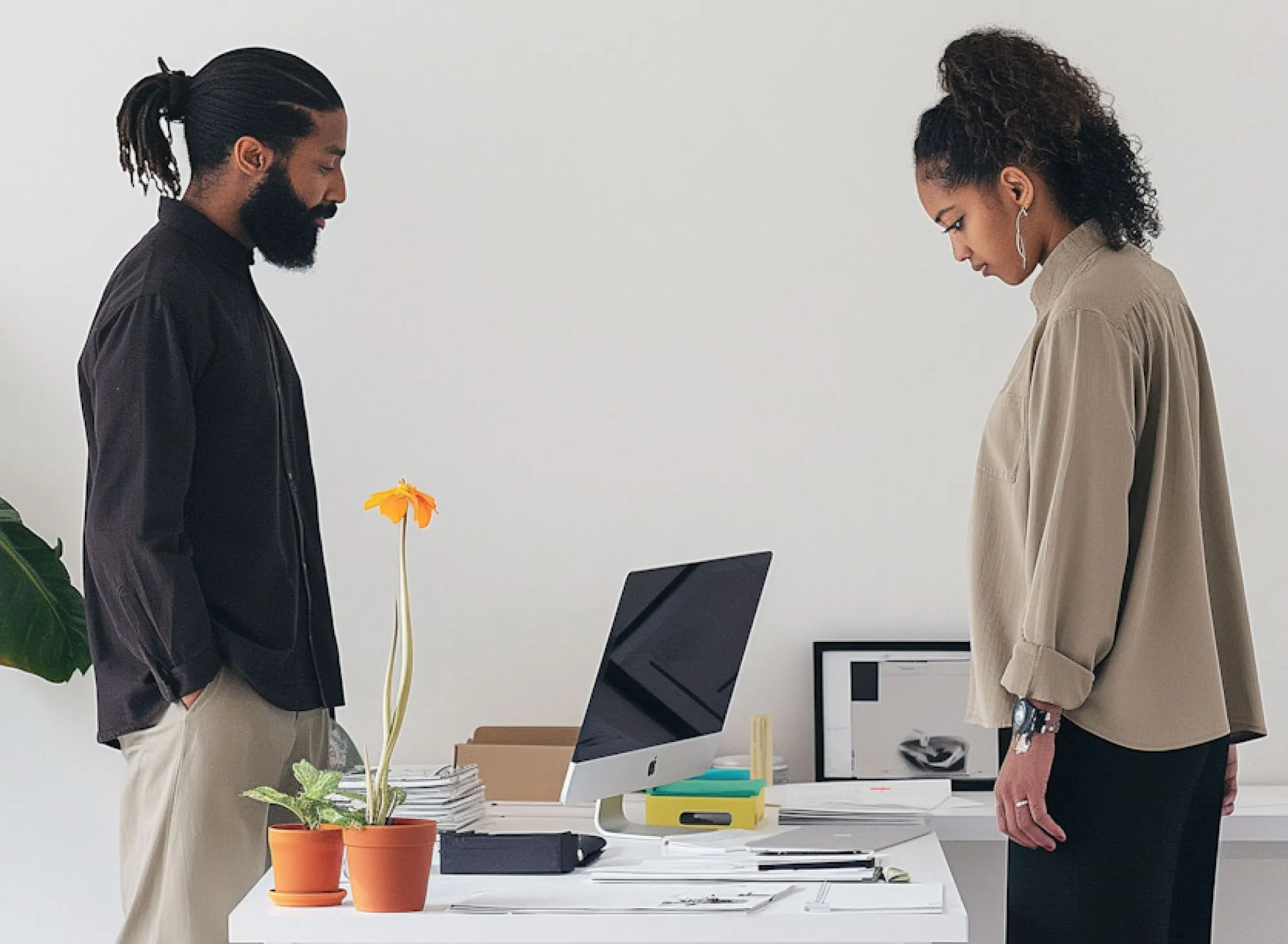 Man and woman standing across from each other at a white desk with an iMac and potted plants, both looking down pensively.