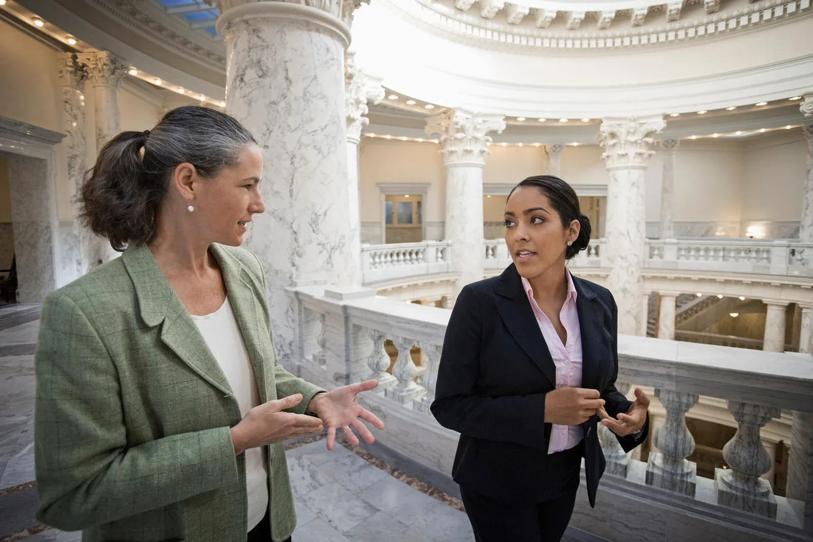 Two professional women in business attire talking inside a government building with marble columns and railings.