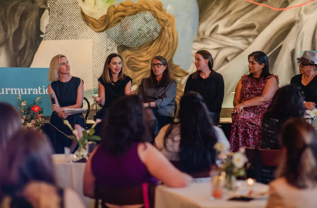 Six women seated on stage in a panel discussion with an audience in the foreground.