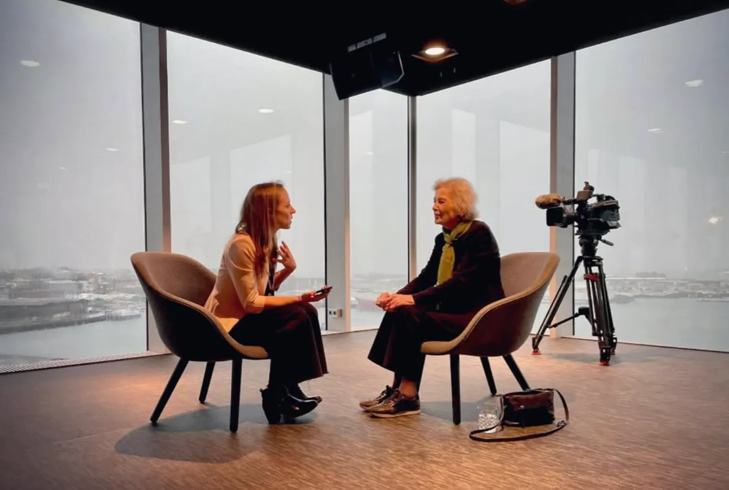 Two women seated in modern chairs facing each other in a high-rise room with large windows, a video camera on a tripod positioned nearby.