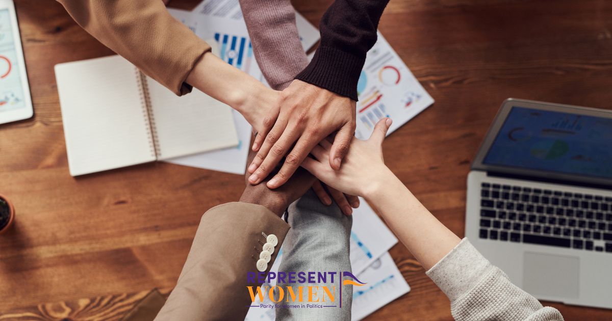 Overhead view of diverse hands stacked together over a wooden desk with charts, a notebook, and a laptop, representing teamwork and collaboration.