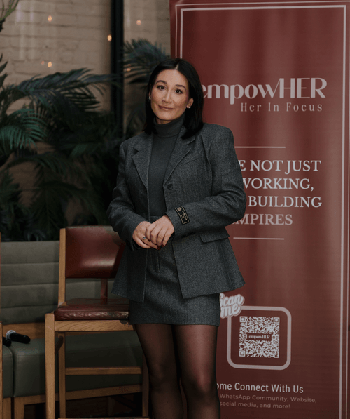 Woman with black hair wearing a gray blazer and skirt standing indoors near a maroon banner with text about empowHER.