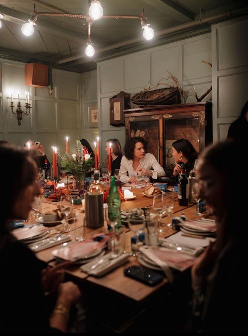 Group of people seated around a wooden dining table with candles, flowers, food, and drinks in a warmly lit room with paneled walls.