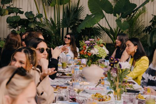 Group of women gathered around a long table with plates of food, drinks, and floral centerpieces in a lush outdoor setting.