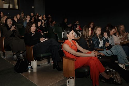 Audience of diverse women seated in dark theater seats attentively watching an event.