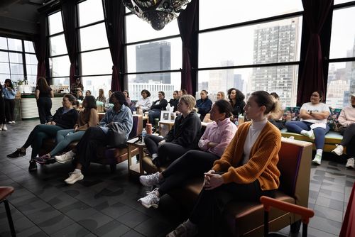 Group of people seated indoors in a modern room with large windows, attentively listening.