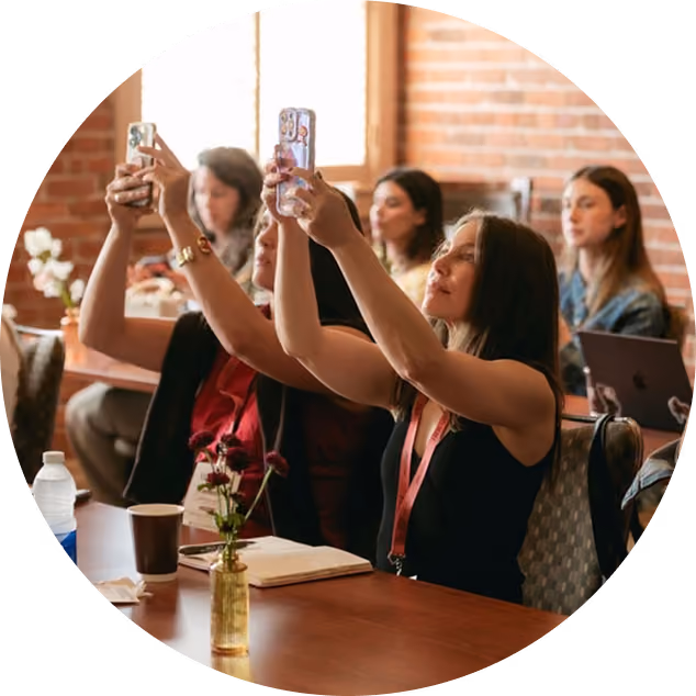 Two women seated indoors at a table, holding their smartphones up to take pictures or videos, with others seated in the background.