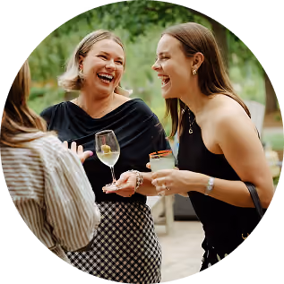 Two women laughing and enjoying drinks outdoors, one holding a glass of white wine and the other a cocktail, with a third person partially visible.