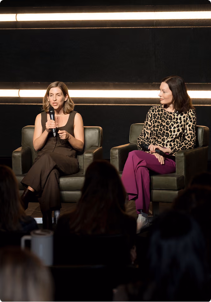 Two women seated in green armchairs on a stage with dark background and horizontal light strips; the woman on the left speaking into a microphone, wearing a sleeveless brown outfit, the woman on the right listening, dressed in a leopard print top and purple pants.