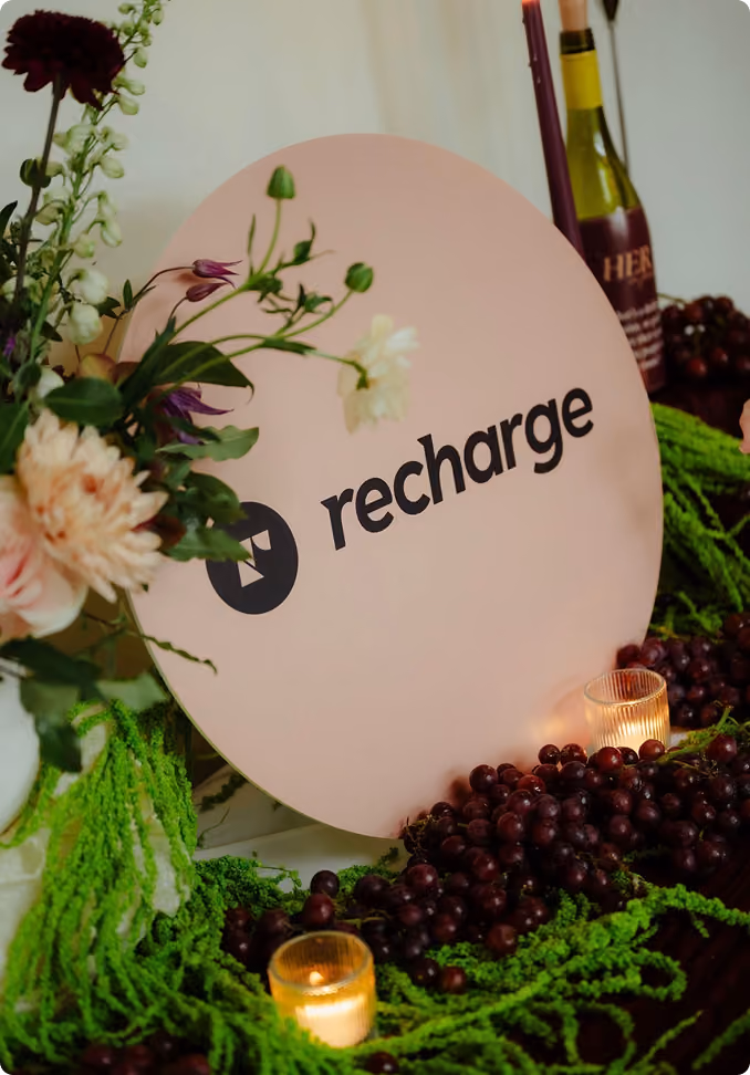 Round pink sign with the word 'recharge' in black letters surrounded by green foliage, flowers, two lit candles, and bunches of dark grapes.