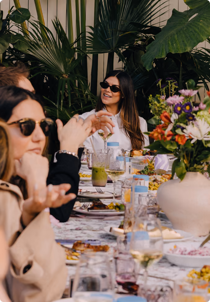 A group of people wearing sunglasses sitting at a table outdoors, sharing a meal with various dishes and drinks, surrounded by large green plants, including a vase with colorful flowers in the foreground.