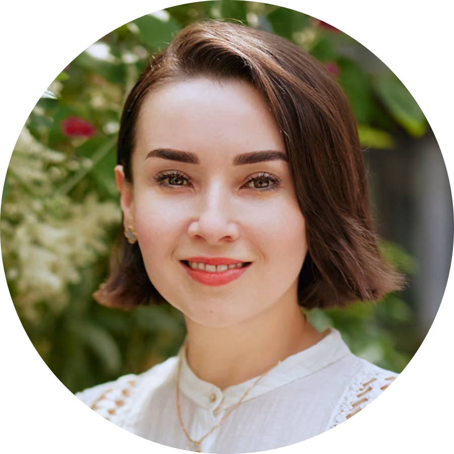 Portrait of a smiling woman with short brown hair and natural makeup wearing a white blouse, with blurred green foliage in the background.