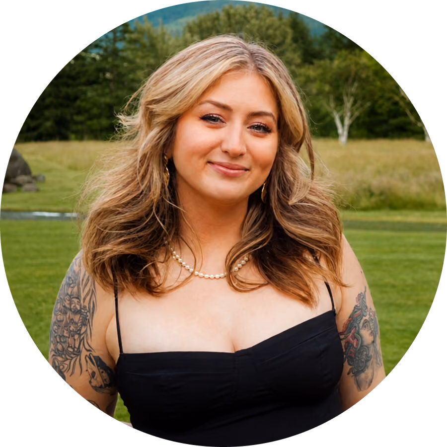 Woman with wavy blonde hair wearing a black sleeveless top and a pearl necklace, standing outdoors in a green grassy area with trees in the background, smiling softly.