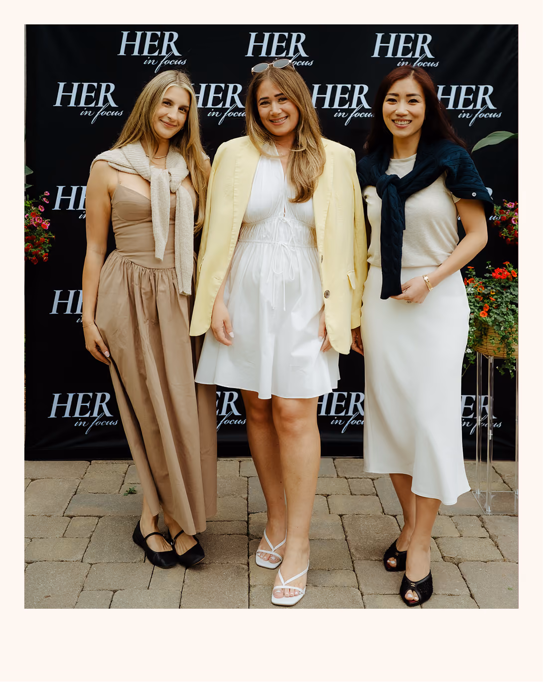 Three women standing on a stone pavement in front of a black backdrop with repeated white text 'HER in focus'. The woman on the left wears a tan strapless dress with a light sweater tied around her shoulders and black shoes. The woman in the middle wears a white dress with a light yellow blazer draped over her shoulders and white sandals, with sunglasses on her head. The woman on the right wears a white sleeveless dress with a dark sweater tied around her shoulders and black open-toe shoes. There are red flowers visible on both sides of the photo.